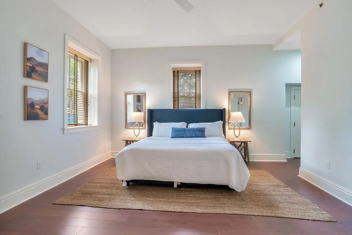 A tranquil bedroom featuring a plush bed adorned with soft linens, framed artwork above the headboard, bedside lamps casting a gentle glow, and a patterned area rug. Natural light filters in through the window, creating a serene, peaceful atmosphere perfect for relaxation.