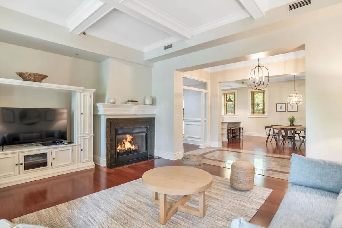 This image features a cozy living room with a central fireplace, a comfortable couch, coffee table, and television. The setting is warmly lit with natural light, and the decor includes tasteful artwork and restored fixtures, setting a historic yet inviting tone.