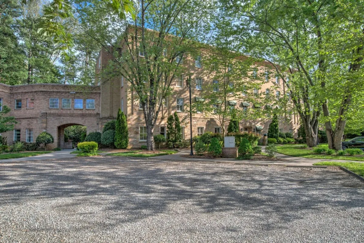 The image showcases the exterior of a historic brick building surrounded by lush greenery and a gravel parking area. Paths and trees create a welcoming environment, perfect for guests to enjoy morning coffee on the patio or gather outside for casual conversation. The overall mood is warm and inviting, suggesting a communal atmosphere for groups.