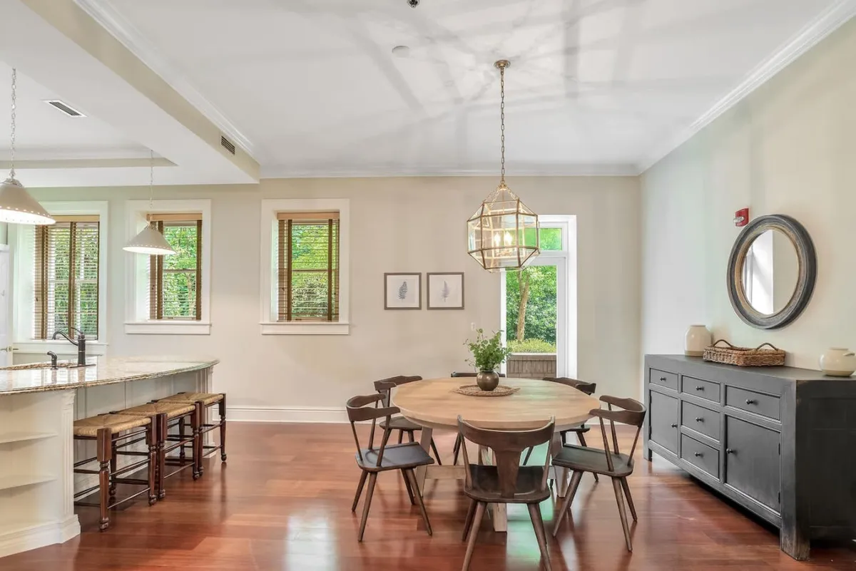 The image centers on a spacious dining area, featuring a large wooden table under a statement chandelier and surrounded by tall windows. The natural light streaming through the windows enhances the cozy ambiance, while the sideboard and tasteful decor evoke a sense of historic charm and comfort.