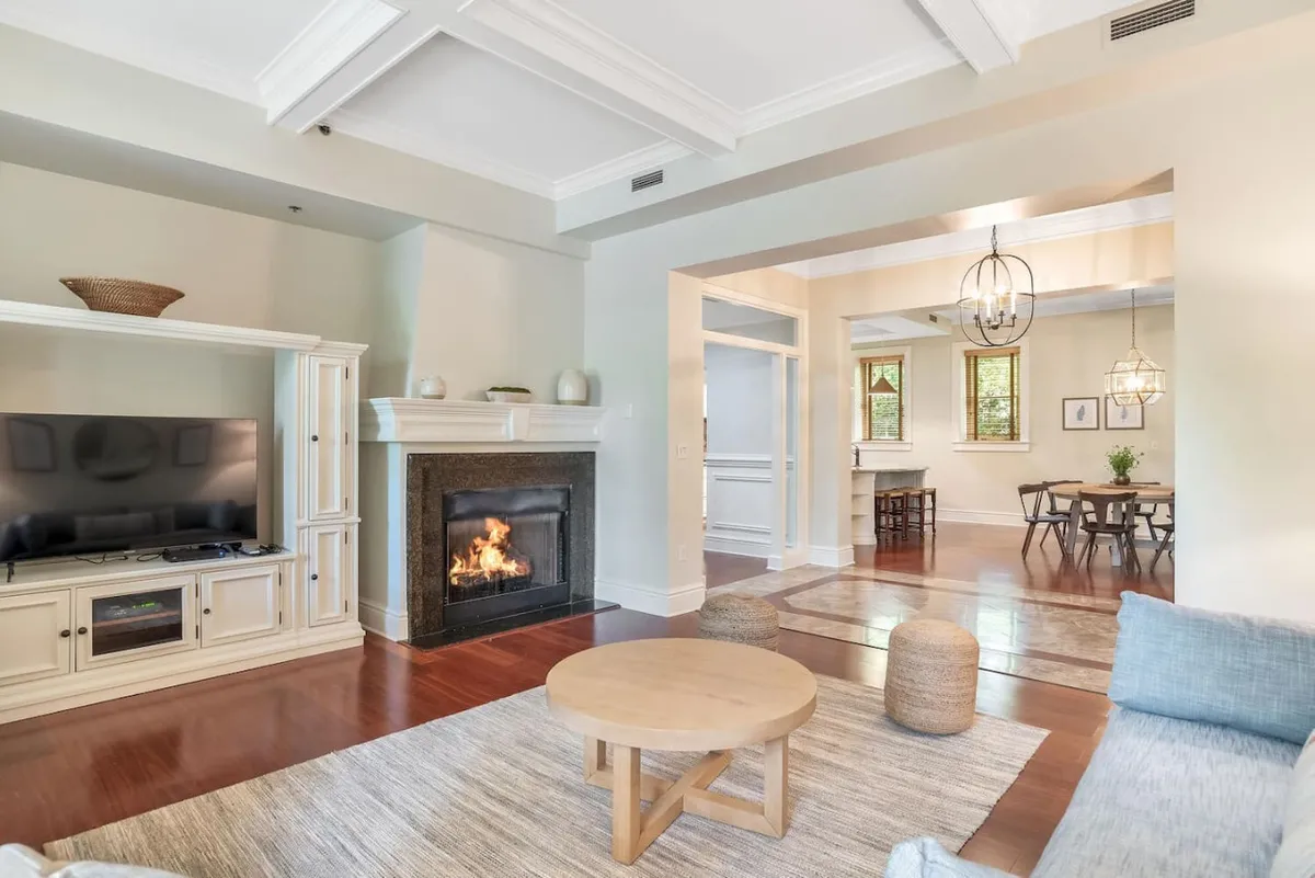 The image showcases a spacious, tastefully renovated living area with a glowing fireplace, comfortable sofa, stylish coffee table, a mounted television, and an inviting dining space—perfect for relaxing or entertaining. The warm lighting and cozy furnishings create a welcoming, home-away-from-home ambiance.
