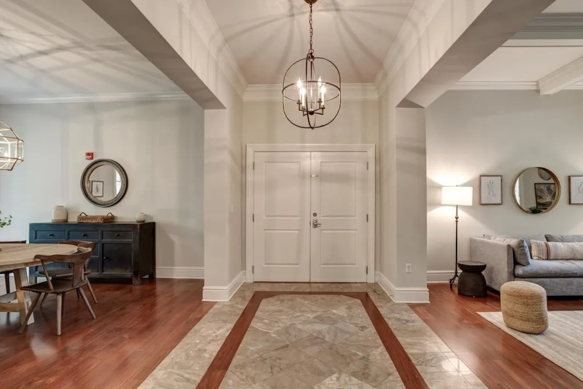 This image offers an inviting view of the home's entrance, flowing directly into the open-plan living and dining area. The scene includes a large dining table perfect for gatherings, a plush sofa for relaxing, and warm decorative elements such as mirrors and a light fixture. Soft natural light filters in, accentuating the hardwood floors and creating a welcoming first impression.