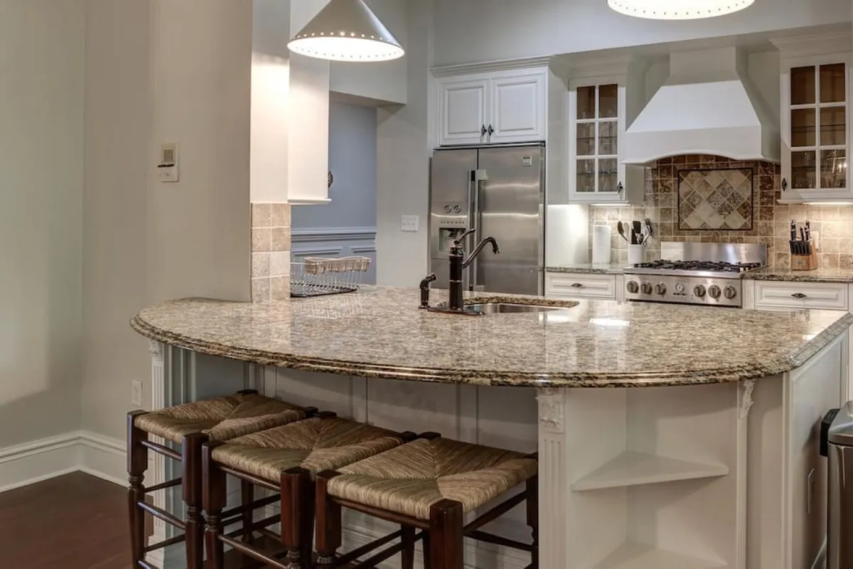This image showcases a beautifully renovated kitchen with a granite countertop, bar stools for casual dining, modern kitchen appliances, and a decorative backsplash. The space exudes warmth and provides ample room for cooking and socializing, with a layout that encourages participation from multiple people.
