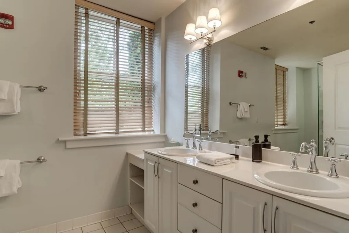 A modern bathroom with double vanity sinks, wide mirrors, a window with blinds for privacy, and soft lighting. The space feels calm and refreshing, ideal for morning routines or relaxing evening preparations.