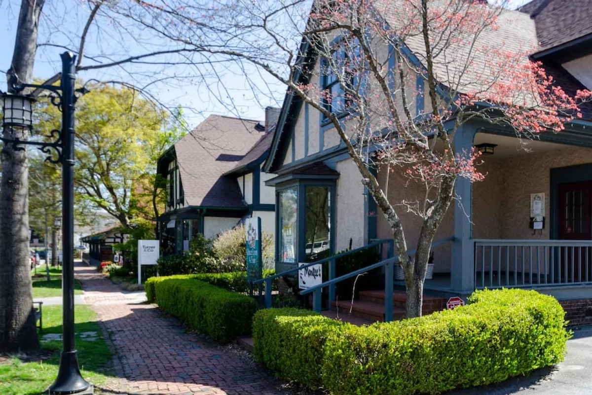 A vibrant neighborhood scene featuring charming houses, manicured hedges, a classic street lamp, and pathways where people are seen strolling and socializing. The image evokes a sense of community and lively local culture, with hints of seasonal decorations and local events.
