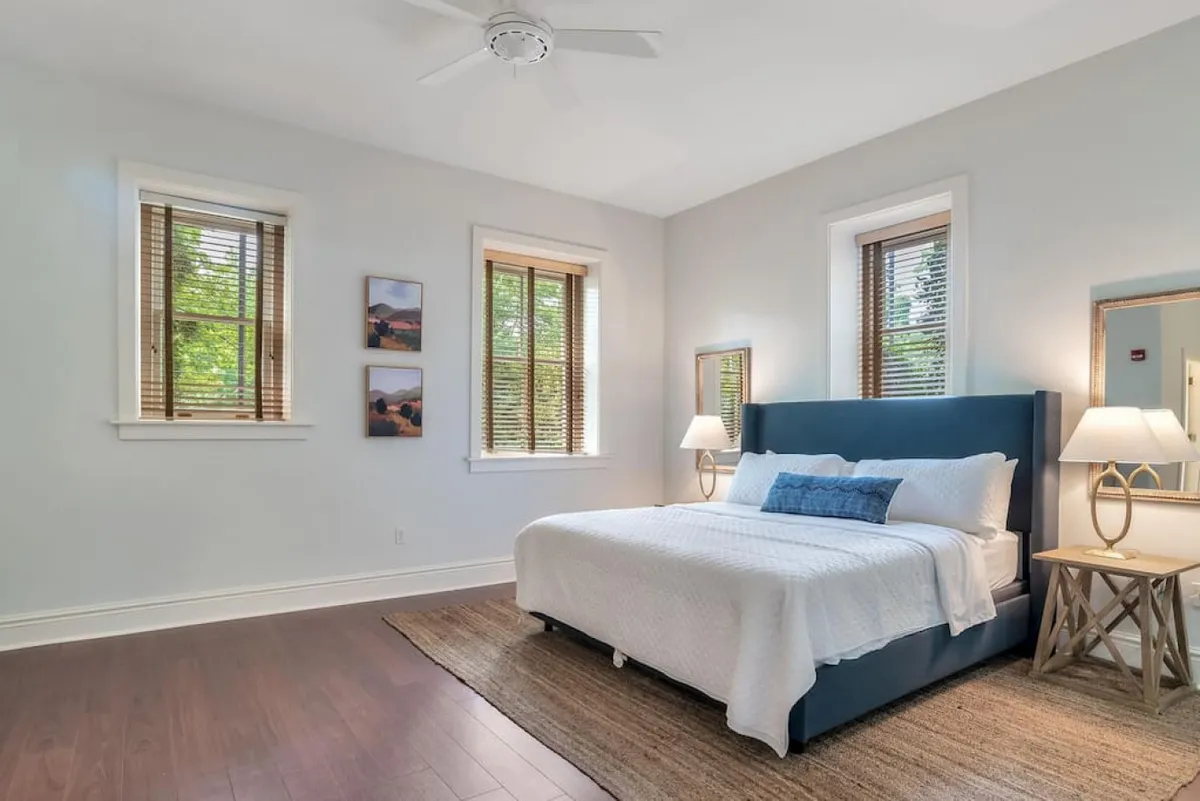 A peaceful bedroom scene with a cozy bed, nightstands on each side, tasteful artwork above the bed, and original windows that bring in daylight. The room combines comfort and a sense of historic charm, ideal for quiet evenings or mornings.