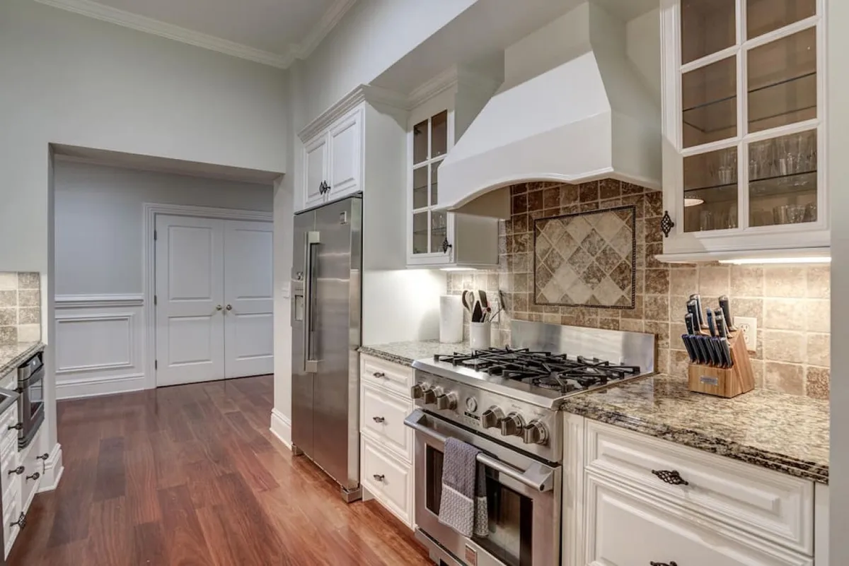 This image highlights the kitchen’s stainless steel appliances, granite countertops, wooden cabinetry, and a decorative tile backsplash. The overall mood is warm and inviting, ideal for gatherings or quiet meals.