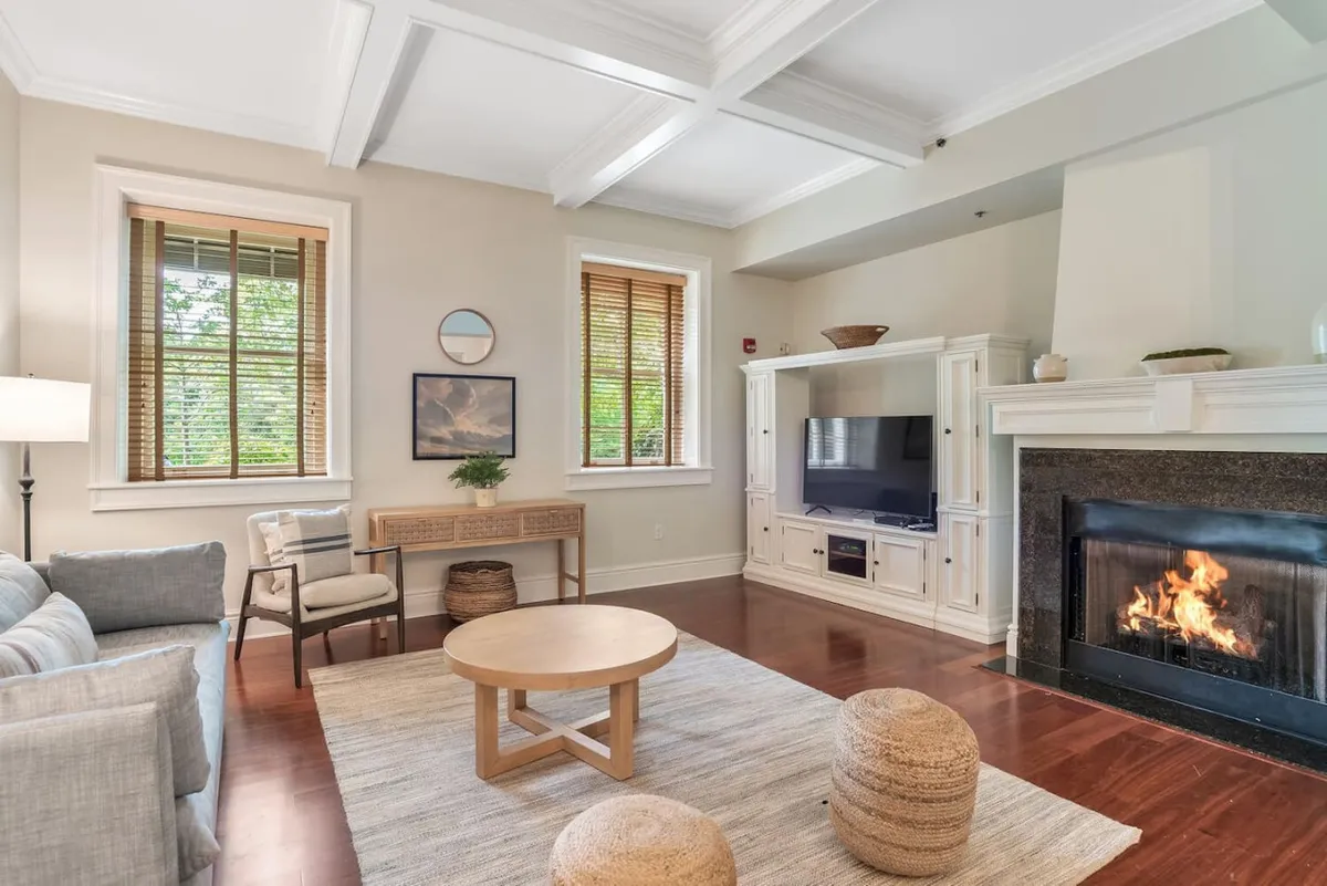 A cozy living room scene featuring a fireplace, comfortable sofa, and coffee table set against a backdrop of artwork and large windows. Sunlight pours in, illuminating the hardwood floors and providing a tranquil, inviting environment for relaxation or conversation.